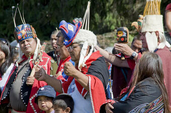 Three men in headdresses of fur, painted carvings and vertical reed-like pieces on top stand in a crowd, two holding carved wooden pieces.