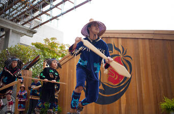 Person in decorated outfit and wide woven hat, holding wooden oar, on one leg mid-dance with 2 dancers behind, and more people standing in background.