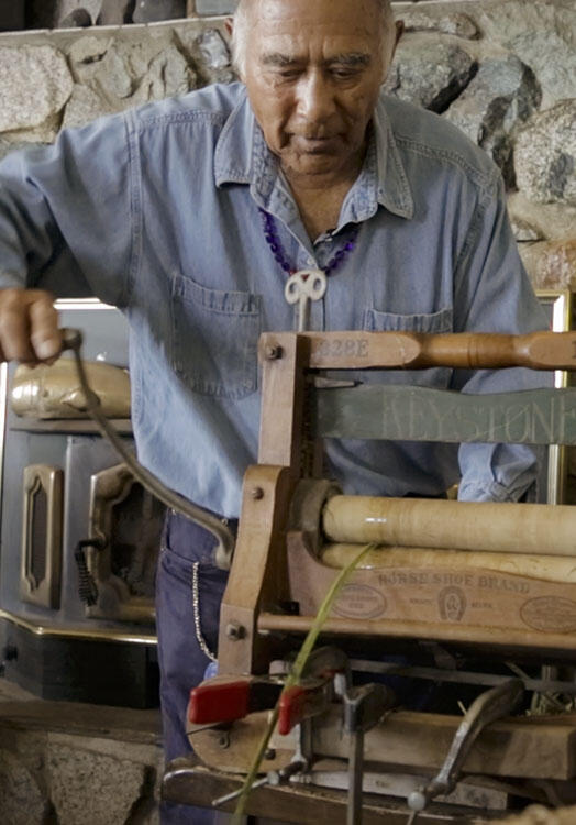 Ed Carriere wears a buttoned shirt and beaded necklace, and uses a wooden, hand-cranked wringer.