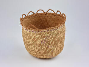 Round woven basket trimmed at the top with protruding woven half-circles in a row on one half and one small handle on the other half. 