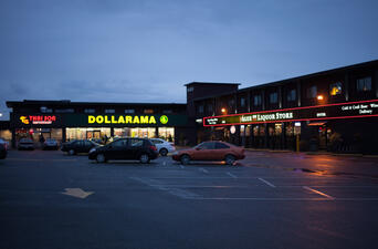 Strip mall at nighttime with (from left to right) Thai Son Restaurant, Dollarama, Value on Liquor Store and a large parking lot with ten parked cars.