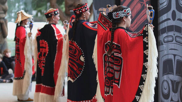 Four women stand next to one another wearing ceremonial dress, and hold carved wooden objects in their hands at arm's length.