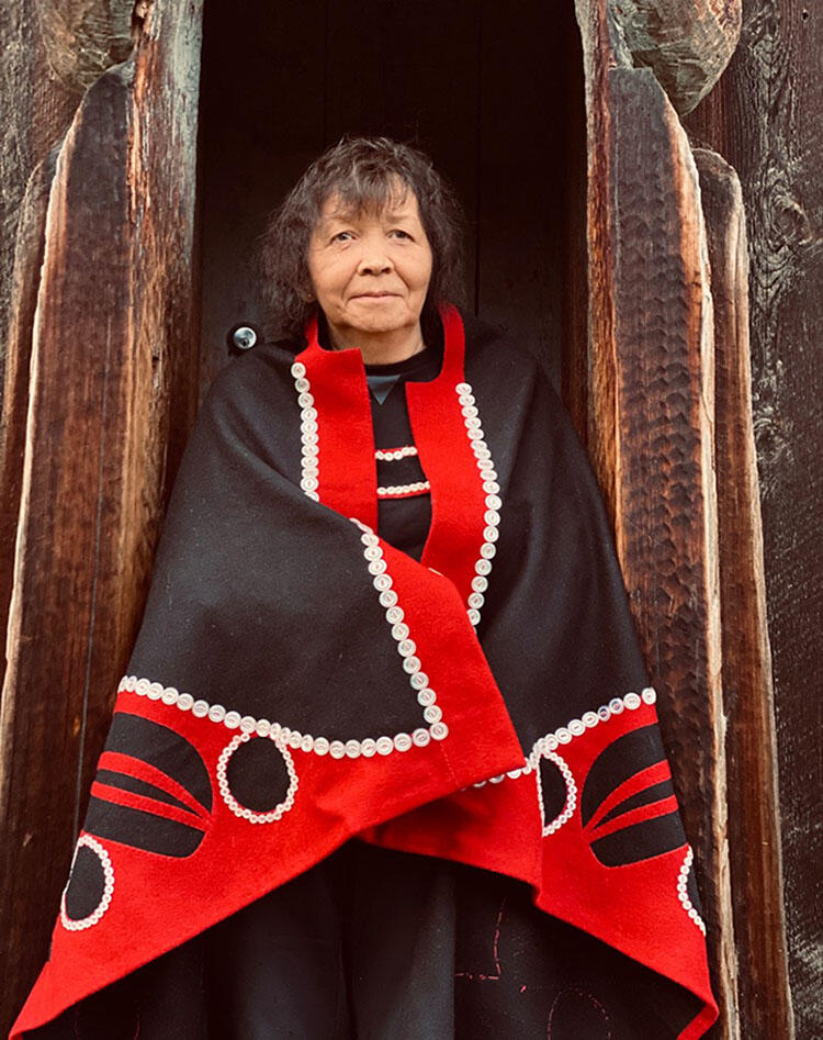 Woman in a dark patterned cloak with colorful trim stands in front of wooden structure.