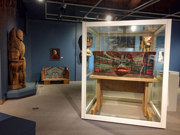 A brightly painted and decorated carved wooden chest sits in a glass cube museum display in a gallery with other wooden carvings and masks on view.