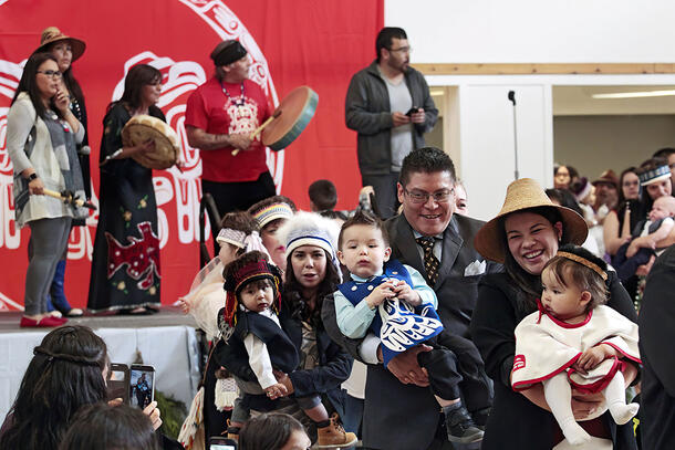 Four people carrying babies as they walk, with performers on a stage behind them holding drums and more people in left foreground, right background.