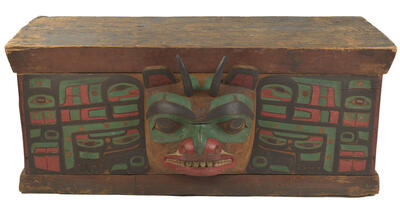 Wooden chest with carved, horned figure at center surrounded by additional carved and painted patterns in multiple colors.