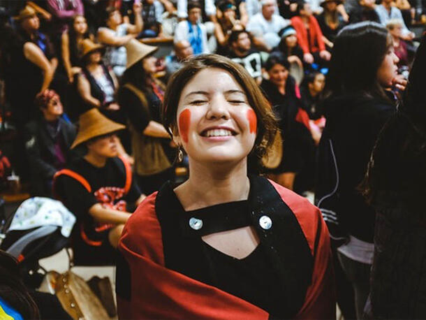 Woman with colorful paint on each cheek smiles with eyes closed with large crowd behind her.