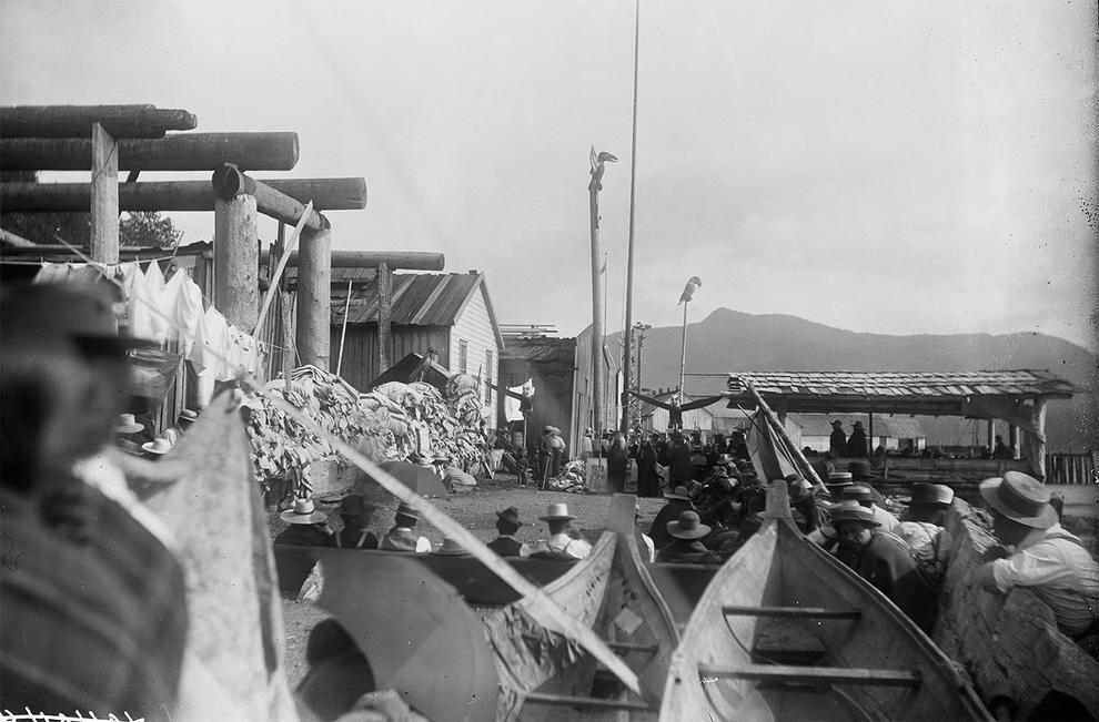 Many people gather along edges of space near wooden buildings and structures and around eagle figure in background, with dinghies in the foreground.
