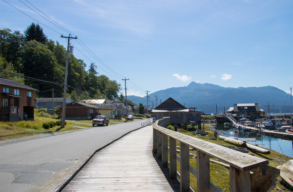 Paved road along coastline with wooden walkway near water, buildings and lawn to the right and a dock and boats to the right.