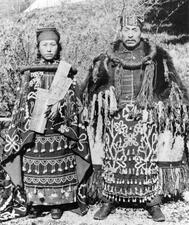 A woman and man stand side-by-side in elaborate tasseled skirts, thick robes, and headpieces.