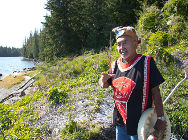 Chief Kwaxitola Henry Seaweed stands on a tree-lined shoreline.