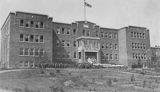 Archival black and white photo of an imposing building from a distance. Dozens of children are lined up outside.