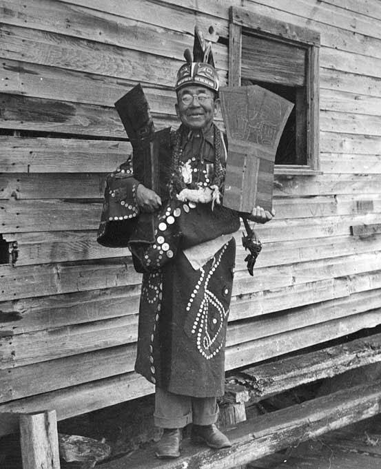 Older man with glasses wearing beaded robe and elaborate headdress holds a metal piece with designs on them in each hand.