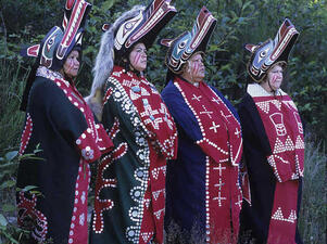 Four women stand in row wearing painted headpieces shaped like stylized wolf heads, beaded robes, with paint on their right cheeks.