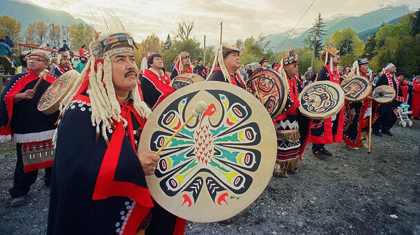 Man in headdress and robe with handheld drum in foreground with row of men in headdresses with drums behind him and more people and trees in the back.