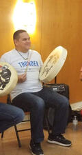 Tsah‘tse-in is seated in a chair and plays a drum.