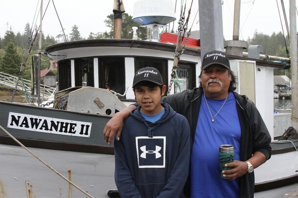 Older man stands with arm around the shoulders of a teenager, posing in front of a fishing boat named Nawanhe II.