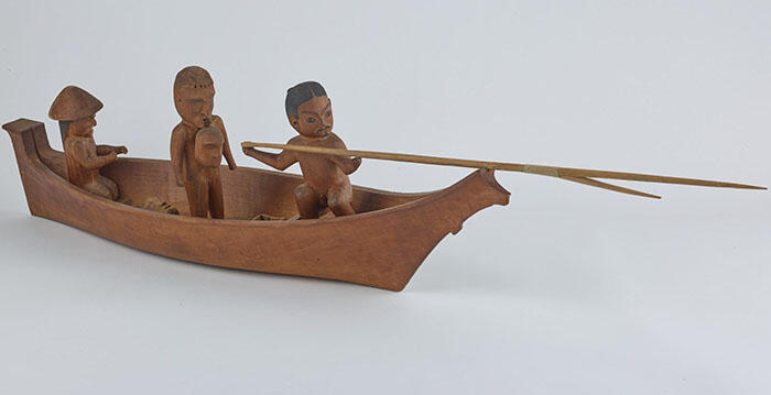 Wooden carving of three figures seated in a canoe; one figure holds a spear.