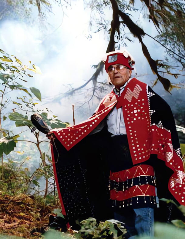 High Chief Nuximlayc stands in a forest clearing wearing traditional garments and holding a carved wooden object.