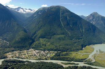 Mountain covered in greenery above a small town and a river. 