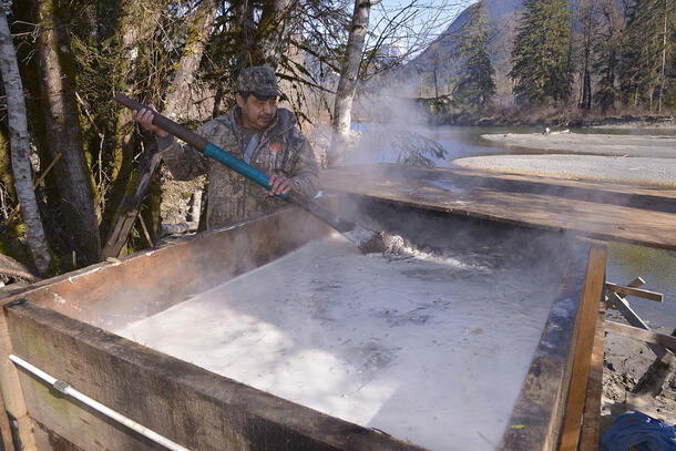 Bert Mack stands next to a large wooden box filled with fish cooking in a broth, and uses a large wooden paddle to stir the fish.