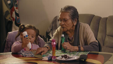 Elderly woman and little girl sit at a table holding bingo daubers over bingo cards, as the older woman helps the child mark her bingo card.