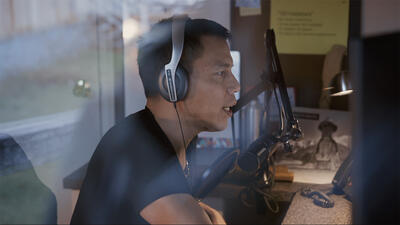 Man wearing over-ear headphones on sits at a desk and speaks into a mic. 