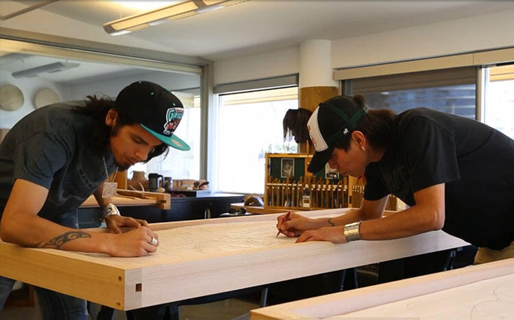 Two students lean over a door-shaped piece of wood, drawing a design on it.