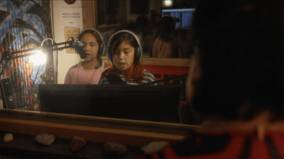 Two kids in a recording room wear over-ear headphones and sing into long microphones. 