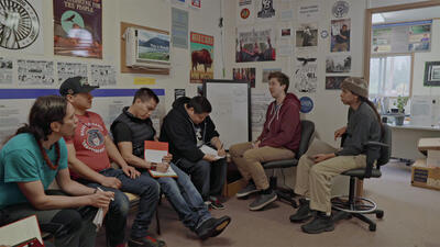 Six casually dressed people sit in a room decorated with various posters, talking to each other while a few take notes.