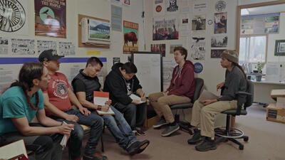 Six casually dressed people sit in a room decorated with various posters, talking to each other while a few take notes.