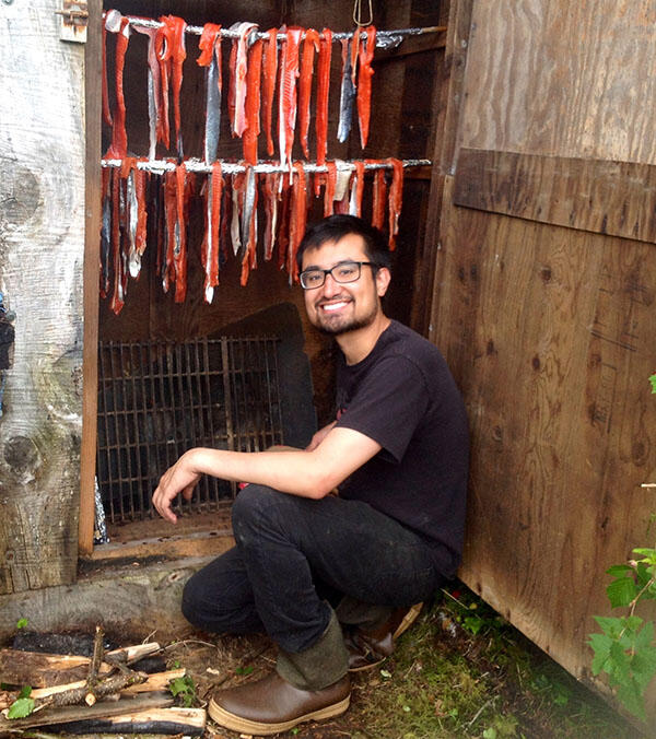 Man sits next to smoker with salmon fillets suspended on racks above it.