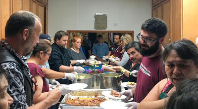 Large group of people stand at a long table filled and serve food.