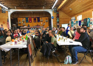 Large groups of people seated at two long tables in a community space.