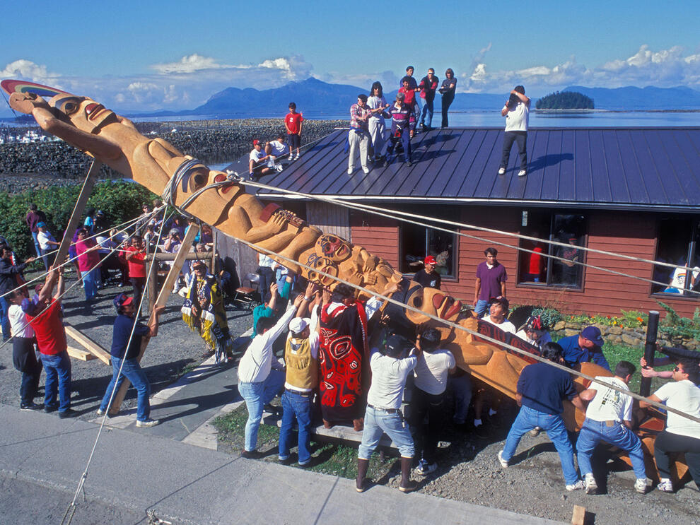 Roughly 20 people help lift carved pole into upright position using rope, wood pieces and hands as small crowd watches, some on nearby rooftop.