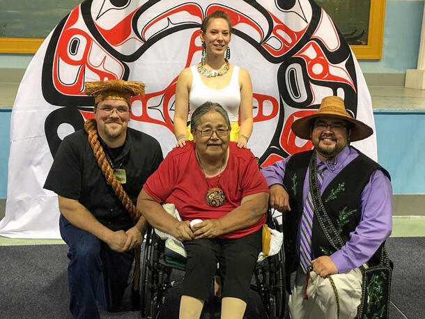 Woman in a wheelchair surrounded by two men on her sides and a woman behind her. Man on left wears straw headpiece and man on right wears large hat.