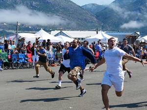 6 men run in a foot race as a crowd of people watch, with mountains in the background.