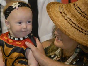 Baby in a floral headband and patterned outfit is held by a woman in a straw hat facing away, as another person's hand reaches for the baby.