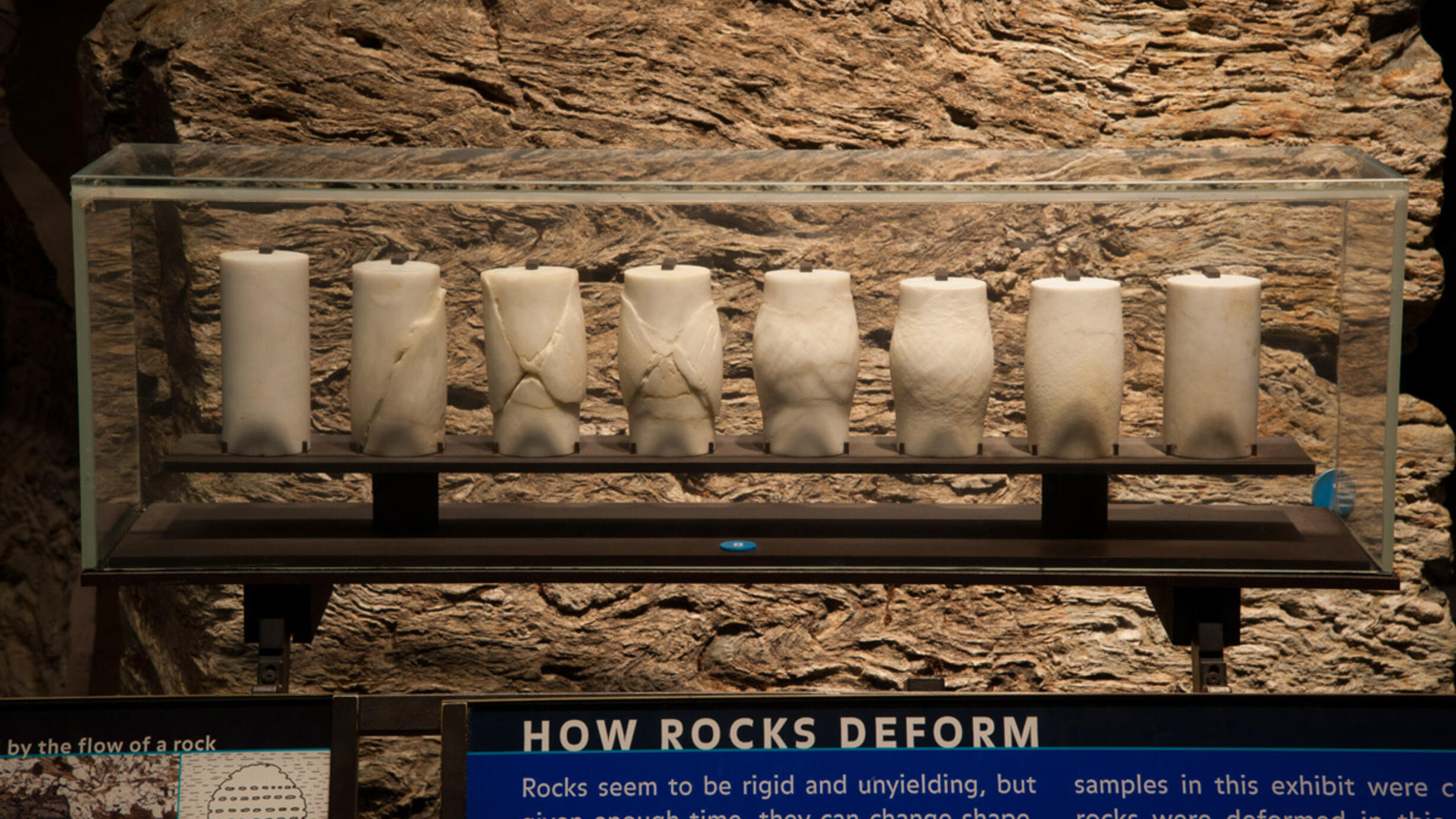 Eight cylinder-shaped samples of marble displayed from left to right in different states of deformation. An explanation panel states how rocks deform under heat and pressure.