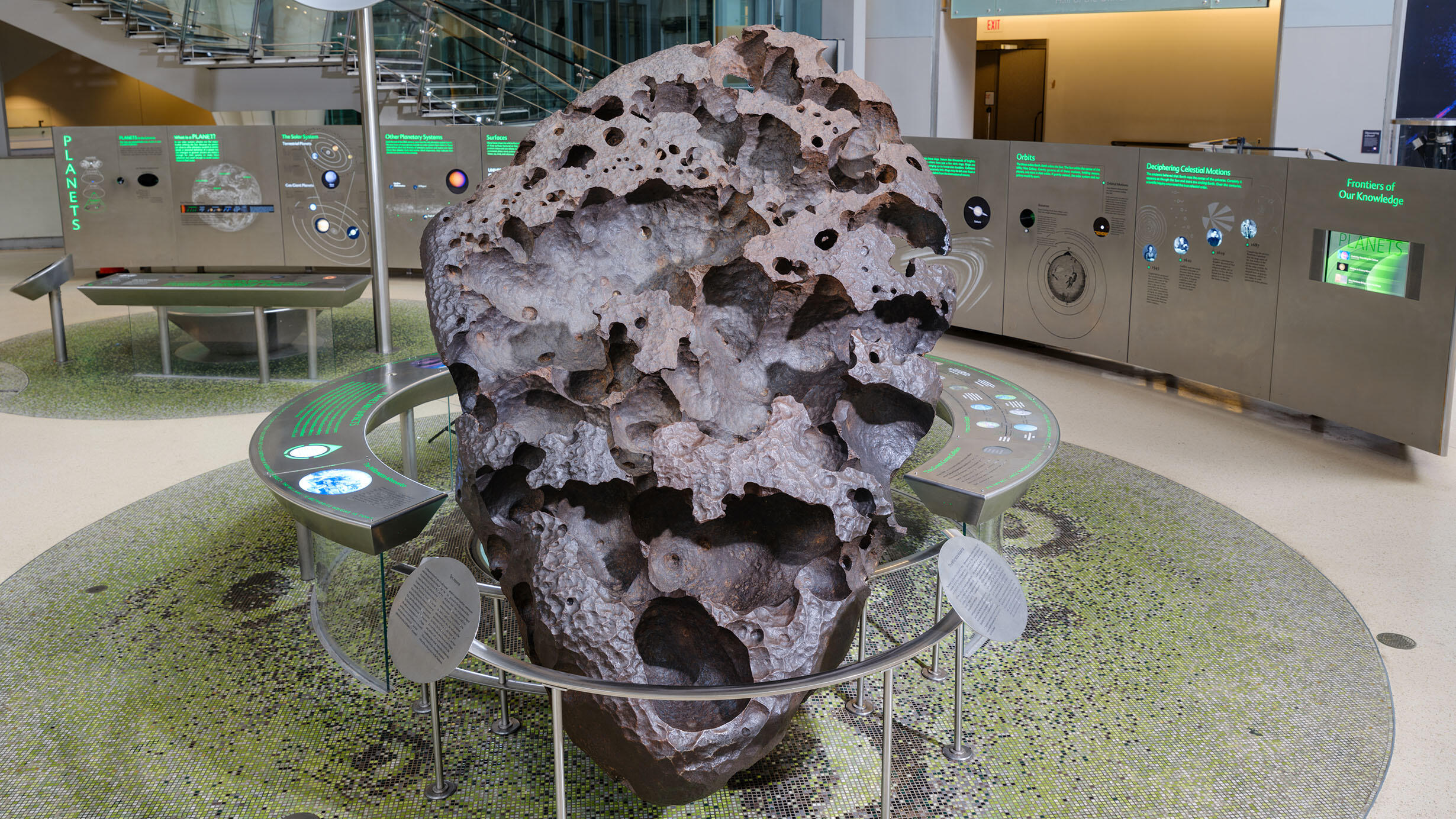 A large, pitted iron meteorite surrounded by a metal barrier with exhibit text on display in the Hall of the Universe.