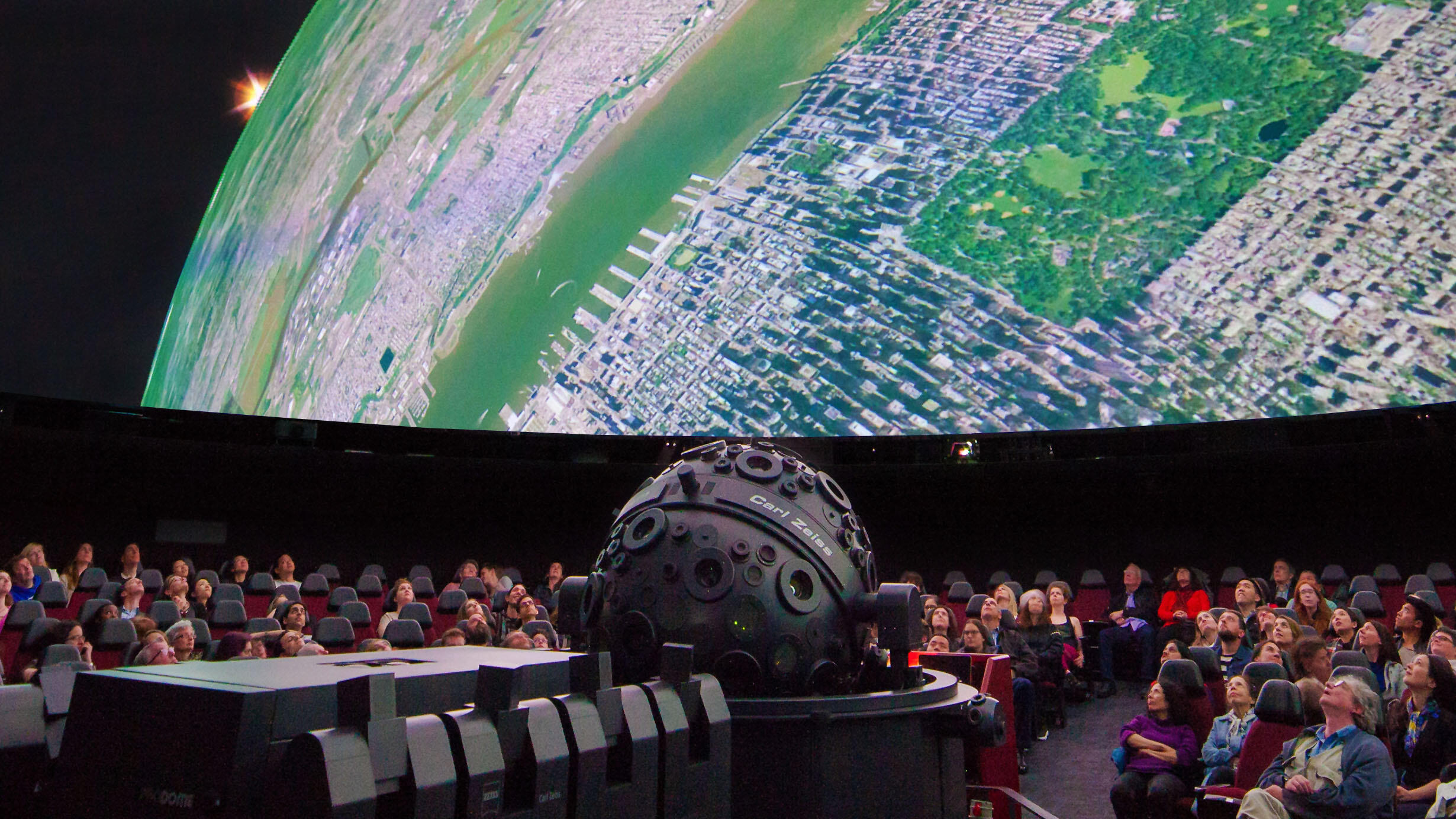 Visitors seated in chairs surrounding the Zeiss projector view images from space on the giant screen inside the Hayden Planetarium.