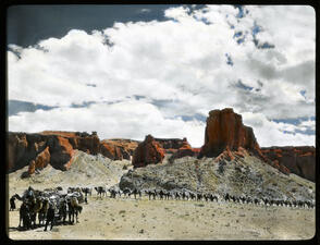 Caravan of camels walking across desert landscape with red cliffs in background.