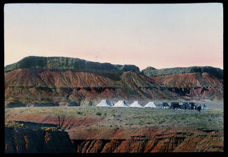 Campsite with white tents and parked black cars in desert against backdrop of red cliffs.