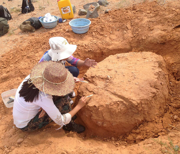 Two people in large hats crouch on red sandy ground to work on red mound.