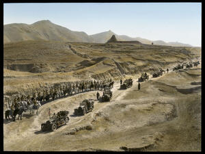 Caravan of black cars drive down the desert road as men on horses wait on the side of the road.