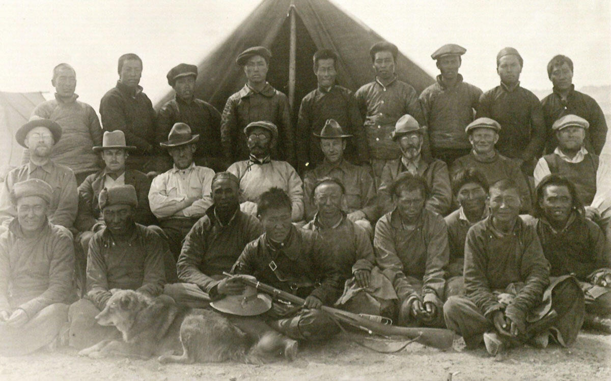 26 men and dog pose in three rows in front of tent.