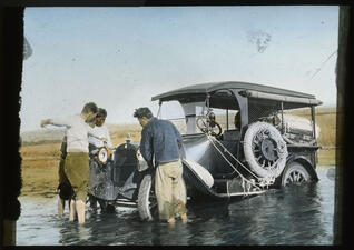 4 men around the front of a black car that has driven into shallow water.