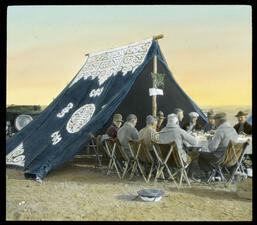 10 men sit around a table partially beneath a blue tent at a desert campsite.