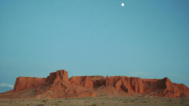 Landscape of red cliffs against large blue sky with small moon.