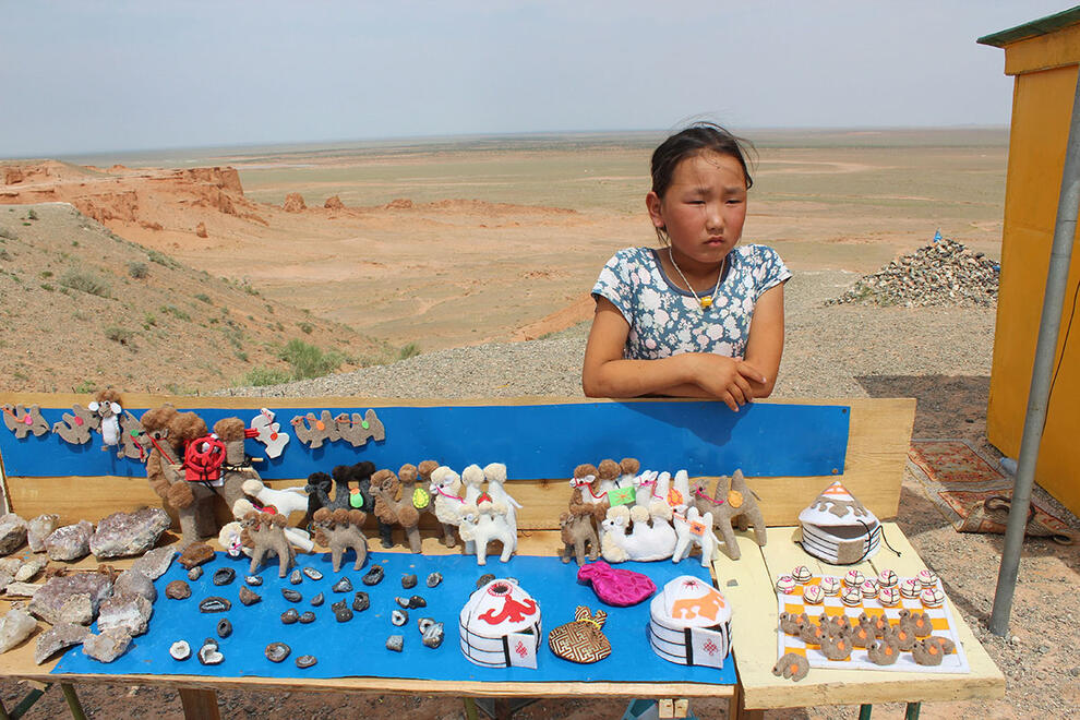 Young girl stands behind stall selling souvenirs in desert setting.